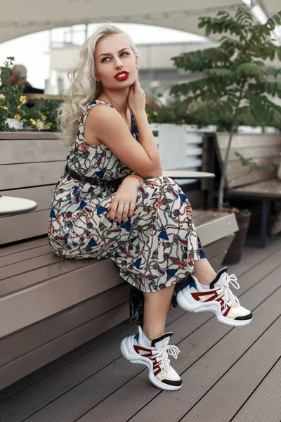 时尚漂亮的年轻女子与红色的嘴唇在时髦的礼服穿着时尚运动鞋坐在街上的木凳上放松 — 图库照片
