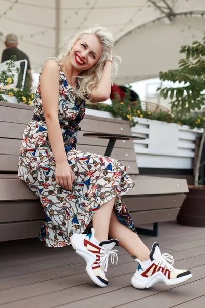 愉快的美丽的女孩在老式的礼服与图案和时髦的运动鞋坐和放松在长椅在城市 — 图库照片