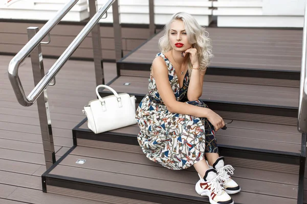 美丽的年轻优雅的模特女士穿着时尚的夏季礼服和运动鞋与白色时尚袋坐在木制楼梯上 — 图库照片