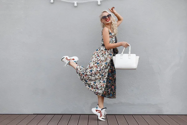 时尚漂亮的时髦模特的女人与太阳镜与一个白色的袋子 穿着图案和时尚的运动鞋跳近灰色的墙壁 快乐购物的生活方式 — 图库照片