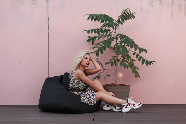 时尚漂亮的年轻模特女士穿着时髦的夏装 鞋子坐在一个装着绿色树的袋子椅上 靠近粉红色的墙 — 图库照片