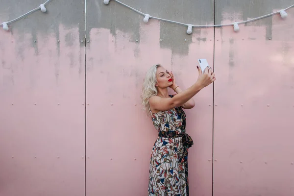 漂亮的年轻女子在时髦的复古礼服采取照片上的街道附近的粉红色墙上的电话 — 图库照片