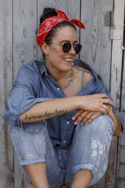 Mutlu güzellik Amerikalı kadın bir kot gömlek ve mavi jeans şık güneş gözlüğü ile bir ahşap duvar oturur. Pinup stili