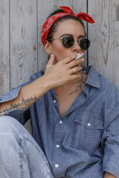 Beautiful young stylish woman with sunglasses and a red bandanna in a denim shirt and vintage jeans sits near a wooden wall and smokes a cigarette