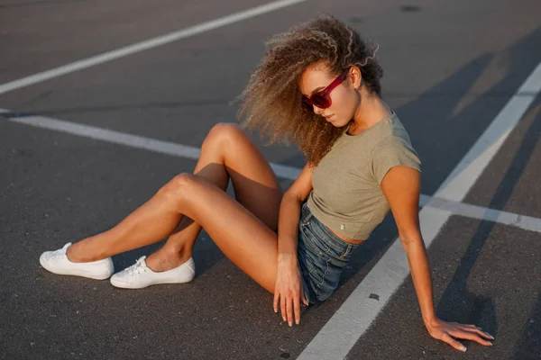 时尚美丽的年轻卷曲的女孩在太阳镜时尚牛仔裤衣服与白色运动鞋坐在人行道上的阳光明媚的一天 — 图库照片