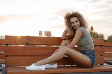 Mutlu güzel kıvırcık olan kadın moda kot şort bir bankta oturan beyaz spor ayakkabı ile