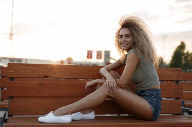 Komik güzel genç şık mutlu kadın vintage kot şort kıvırcık saçlı bankta oturuyor