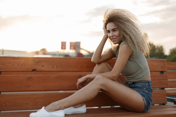 时尚漂亮的年轻女孩卷曲的头发在时髦的牛仔服装与性感的腿坐在板凳上的街道上 — 图库照片