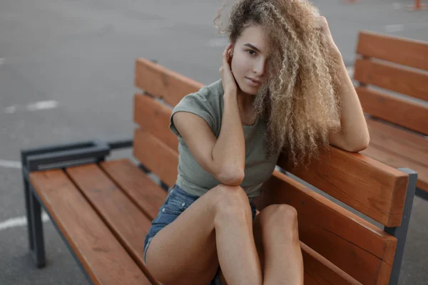 木製のベンチに座っているスタイリッシュなデニムのショート パンツでセクシーな美脚とおしゃれなかなり若いカーリー女の子 — ストック写真