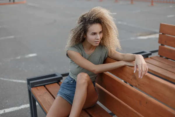 木製のベンチに座っているおしゃれなデニムの服でカーリーの若いスタイリッシュな女性 — ストック写真