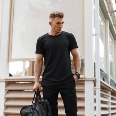 Şık yakışıklı genç Amerikan hipster modeli erkek saç modeli moda olan siyah tişört ile çanta Ahşap Bina cadde üzerinde duran