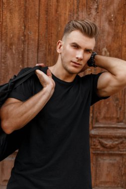 Yakışıklı adamla şık bir saç modeli siyah T-shirt siyah bir çanta ile antika ahşap kapının yanında duruyor.