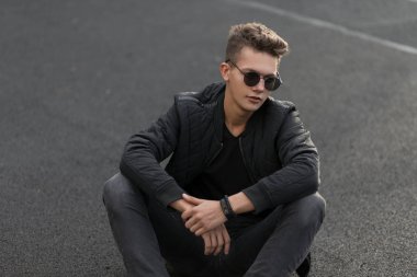Şık yakışıklı genç siyah moda ceket ve siyah kot moda güneş gözlüğü ile sokakta oturuyor