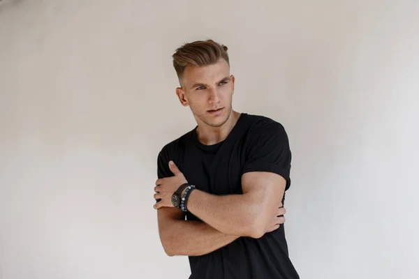 Fashionable handsome young man with hairstyle in a black t-shirt with a watch poses near a white wall on the street