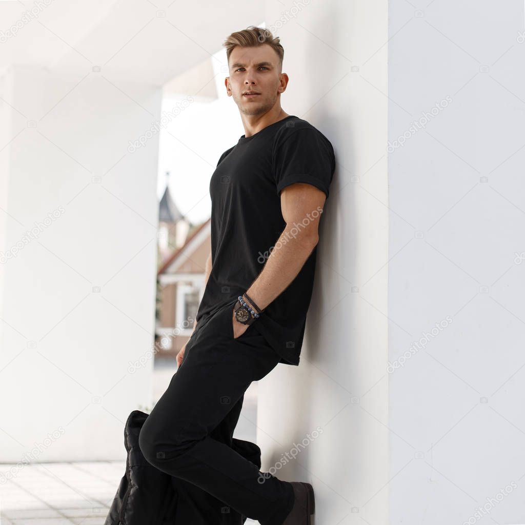 Young fashion model man with bag in black shirt and pants with black bag stands near white wall