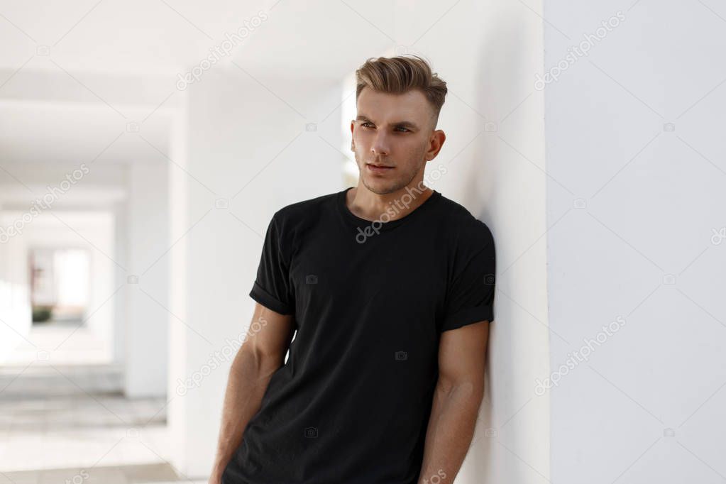 Handsome young american stylish man with hairstyle in fashion black shirt standing near white wall