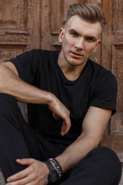 Şık yakışıklı genç bir vintage ahşap kapının yanında sokakta oturan siyah moda giysiler içinde saç modeli ile
