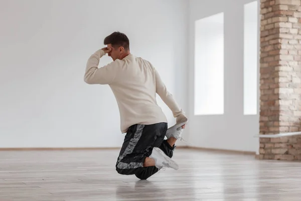 A young man dancing break dance standing on his foot and holding his head in a modern studio, against the background of white walls.