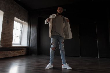 Şık yakışıklı delikanlı ile karanlık bir stüdyoda poz spor ayakkabıları ile mavi sökük modaya uygun kot pantolon moda beyaz ceketli