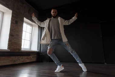 Şık yakışıklı genç çekici erkek dansçı ile moda trendy ceket kot ve beyaz ayakkabılarla dans stüdyosunda koyu arka plan üzerinde dans sökük. Yaşam tarzı dans