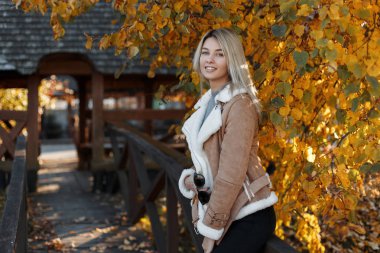 Mutlu genç Şirin ve şık sarışın kız mavi gözleri ve tatlı bir gülümsemeyle sonbahar günü sıcak bir parkta büyük boy sıcak moda giysiler giriyor. Neşeli kadın.