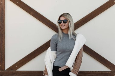 Genç çekici gülümseyen hipster kadın şık deri sonbahar ceketli siyah şık güneş beyaz duvara yakın duruyor. Kız mutlu moda