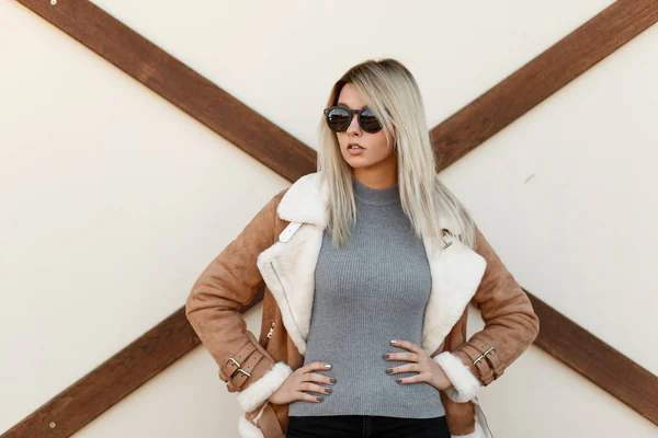 灰色で若いセクシーな驚くほど流行に敏感な女性ニット セーター ジャケット黒サングラスをかけた屋外木造軸組と白い壁の近くでウエストに保持手でポーズします 現代のトレンディな女の子 — ストック写真