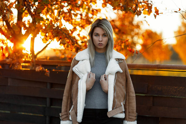 Young pretty fashion girl in a gray knitted vintage sweater and stylish jacket in a park near a wooden fence on the background of golden trees and bright sunlight. Autumn stylish blond woman at sunset