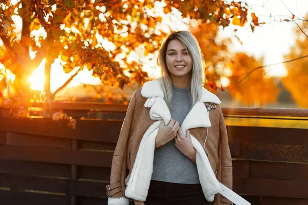 年轻快乐的女人穿着针织复古毛衣和时尚夹克走在公园附近的一个木制的老栅栏在金树的背景下 在明亮的阳光下 秋天愉快微笑的女孩在日落 — 图库照片