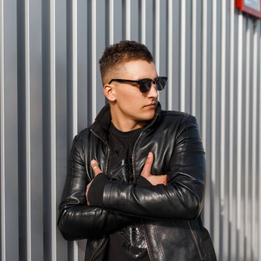 Moda yakışıklı genç bir siyah deri ceket ve trendy kazak içinde şık güneş gözlüğü ile metal duvara yakın duruyor