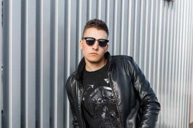 Siyah şık deri ceket ve siyah kazak gözlüklü şık yakışıklı hipster adam metal duvara yakın duruyor