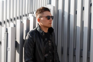 Bir kazak ile şık siyah deri ceketli gözlüklü moda hipster adam metal duvara yakın duruyor