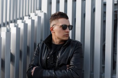 Çekici genç hipster adam siyah güneş gözlüğü ve bir saç modeli sonbahar moda siyah deri ceketle gümüş metal duvara yakın duruyor. Şık modern adam.