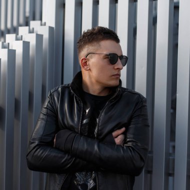 çekici kendine güvenen genç hipster adam siyah güneş gözlüğü ve sonbahar moda siyah deri ceket bir gümüş metal duvara yakın duruyor. Şık modern adam.