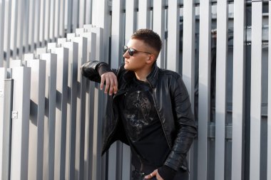Çekici genç hippi erkek saç siyah güneş gözlüğü, trendy siyah ceket ve siyah kazak modeli ile gümüş metal duvara yakın poz veriyor. Şık modern adam. Sunny sonbahar günü