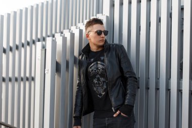 Genç yakışıklı hipster erkeklerde şık bir sweatshirt içinde güneş gözlüğü siyah, son moda deri ceket ve siyah kot güneşli bir günde bir metal duvara yakın duruyor. Çekici şık adam. City Style