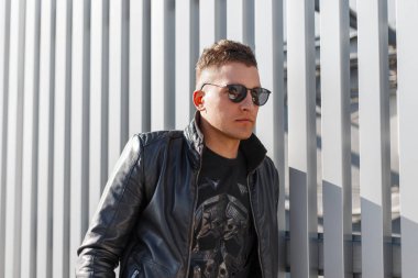 Yakışıklı genç hippi moda adam siyah kazak ve siyah şık ceket şık güneş gözlüğü gri bir metal duvara yakın güneşli bir günde teşkil etmektedir. Çekici moda adam
