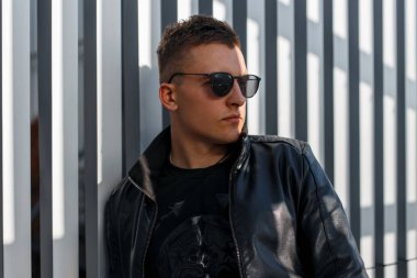 Yakışıklı hipster genç adam siyah güneş gözlüğü ve siyah şık deri ceket gri bir metal duvara yakın güneşli bir günde duruyor. Saç modeli ile çekici şık adam