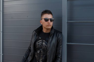 Yakışıklı hipster genç adam siyah şık güneş ve siyah trendy kıyafetler model ve kazak bir metal gri duvara yakın poz moda. Çekici kentsel moda guy
