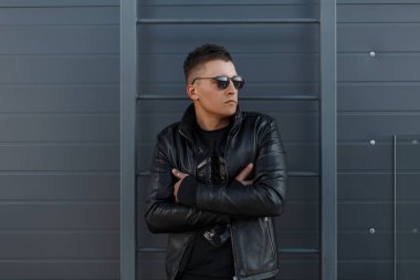 Genç hippi erkek saç modeli güneş gözlüğü siyah şık ceket ve siyah kazak ile gri metal duvara yakın poz veriyor. Yakışıklı olan şık sonbahar siyah giysili