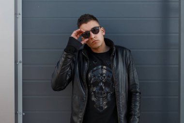 Çekici genç hippi erkek güneş gözlüğü, kafatası desen ve şık siyah deri ceket, siyah kazak metal bir duvara yakın poz ve kameraya doğru görünüyor. Kentsel şık adam