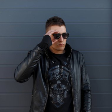 Güneş gözlüğü, kafatası desen ve şık siyah deri ceket gri bir metal duvara yakın poz ile siyah sweatshirt moda genç hipster adam. Kentsel şık adam
