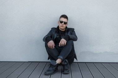 Gri bir duvar yakınındaki bir ahşap zemin üzerinde siyah güneş gözlüğü, siyah deri ceket, şık kazak ve modaya uygun kot pantolon çekici genç hipster adam oturur. Yakışıklı adam