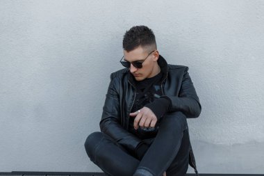 Siyah güneş gözlüğü ve oturma ve gri bir duvara yakın seyir siyah şık sonbahar kıyafetleri yakışıklı şık genç hipster adam. Çekici şık adam.