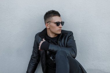 Siyah güneş gözlüğü, siyah ceket, şık kazak ve modaya uygun kot pantolon çekici genç hipster adam açık havada gri bir duvara teşkil etmektedir. Yakışıklı şık modern adam