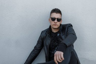Yakışıklı genç hipster adam siyah güneş gözlüğü, siyah ceket, şık kazak ve modaya uygun kot pantolon açık havada gri bir duvara yakın oturuyor. Serin moda adam