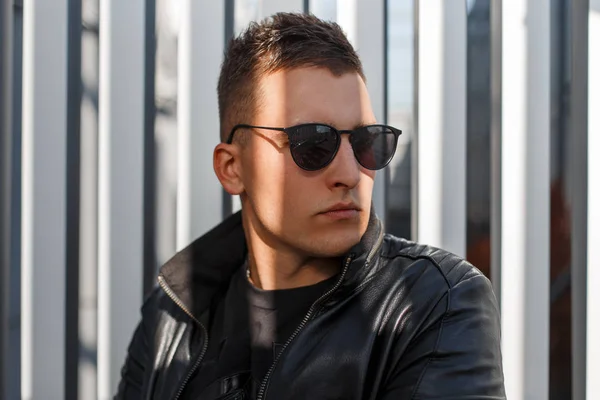 Portrait of an attractive stylish young hipster guy in dark sunglasses and a black leather jacket against a gray metal wall on a sunny autumn day. Stylish man. Close-up