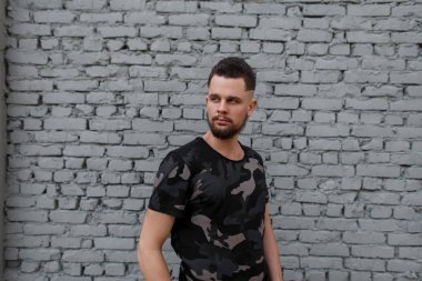 Şık bir kamuflaj çekici acımasız genç adam bir saç modeli ve şık sakal ile askeri T-shirt gri tuğla duvar cadde üzerinde duruyor. Şık adam.