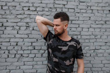 Şık kamuflaj askeri yakışıklı çekici bir genç adam gri T-shirt bir şık saç ve sakal, gri tuğla duvara yakın poz. Moda kentsel guy.