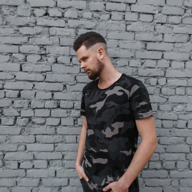 Çekici genç yakışıklı bir saç modeli ve şık sakal ile şık bir askeri T-shirt beyaz duvara yakınında sokakta duruyor. Şık adam.